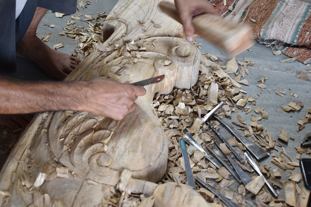 An artisan hand-carving a solid wood furniture panel with chisels and a mallet in the workshop